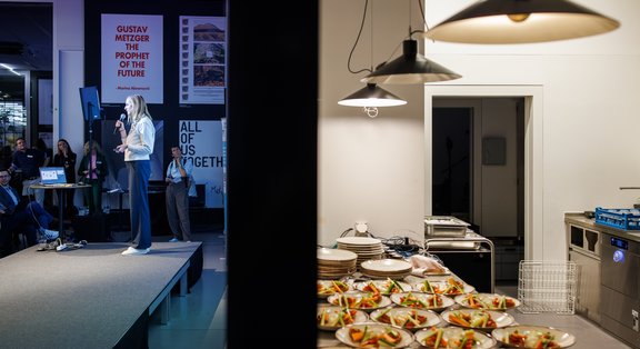 Une personne se tient sur une petite scène et s'adresse à un public. À gauche de l'image, on voit des spectateurs, à droite, une pièce attenante avec de nombreuses assiettes préparées sur un plan de travail.